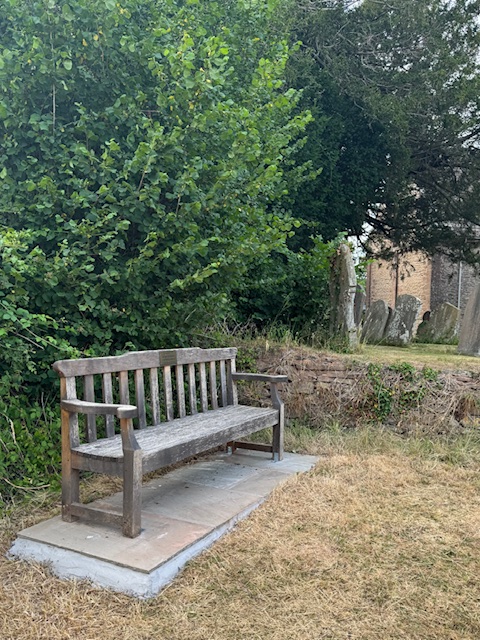 Photo of Photo 1: Wooden memorial bench installed on a stone base, with greenery in the background. Photo 2: Angled view of the memorial bench near a churchyard, surrounded by trees and gravestones. Photo 3: Close-up of plaque on bench that reads: "In memory of George Meadmore, a lifelong resident of Kilpeck Parish."